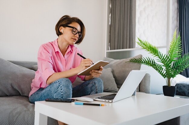 Mulher séria e ocupada com camisa rosa sentada concentrada, fazendo anotações, pagando contas no sofá em casa, à mesa, trabalhando online no laptop de casa