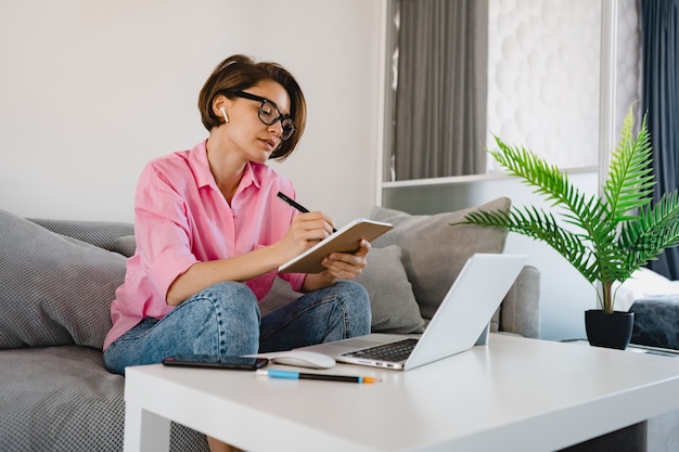 Mulher séria e ocupada com camisa rosa sentada concentrada, fazendo anotações, pagando contas no sofá em casa, à mesa, trabalhando online no laptop de casa