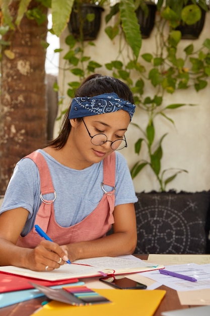 Mulher séria, mestiça, com faixa na cabeça, vestida com camiseta casual e macacão, escreve os registros no bloco de notas