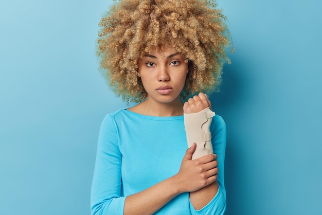 Foto grátis mulher séria tem fratura de braço usa bandagem elástica olha diretamente para a câmera vestida com jumper casual isolado sobre fundo azul paciente do sexo feminino vem na visita do cirurgião ou traumatologista