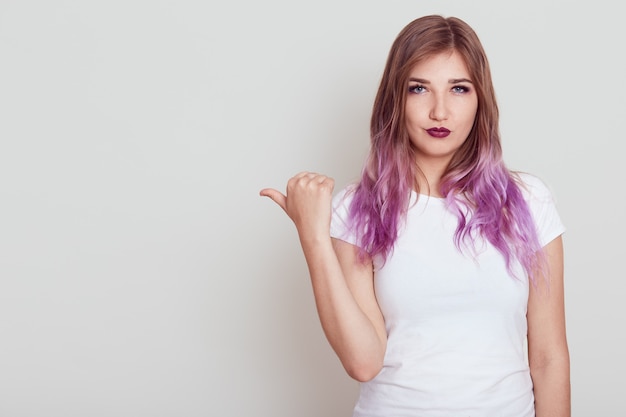 Mulher séria, vestindo camiseta branca casual com expressão estrita, apontando o polegar de lado, mostrando o espaço livre para propaganda, posando isolado sobre uma parede cinza.