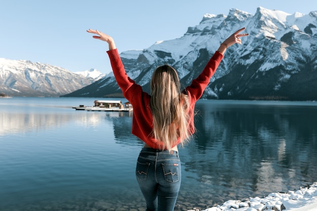 Mulher sexy com corpo magro perfeito em pé na praia perto do lago de inverno. Neve branca no chão e nos picos das montanhas. Cabelo loiro comprido deitado na parte de trás do suéter vermelho.