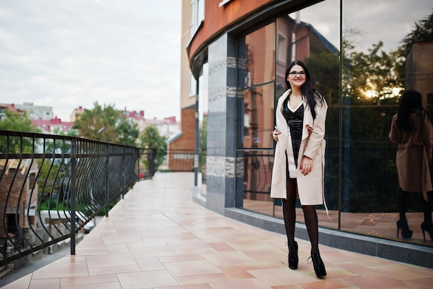 Mulher sexy de cabelo preto em óculos e casaco posou contra prédio com janelas modernas