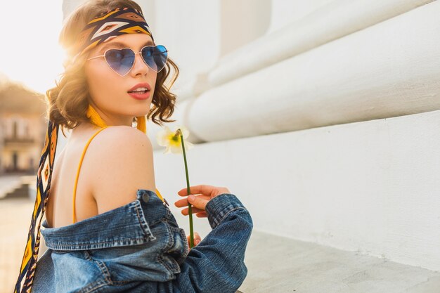 Mulher sexy e atraente elegante em um vestido amarelo elegante usando jaqueta jeans, roupa da moda, tendência da moda primavera-verão, sol, óculos de sol azuis, moda de rua, estilo hippie, acessórios de moda