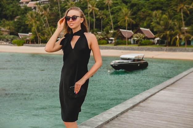 Mulher sexy e atraente luxuosa vestida de vestido preto, posando no cais em um hotel resort de luxo, usando óculos escuros, férias de verão, praia tropical