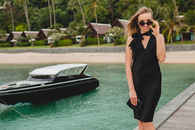 Mulher sexy e atraente luxuosa vestida de vestido preto, posando no cais em um hotel resort de luxo, usando óculos escuros, férias de verão, praia tropical