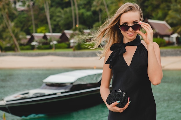 Foto grátis mulher sexy e atraente luxuosa vestida de vestido preto, posando no cais em um hotel resort de luxo, usando óculos escuros, férias de verão, praia tropical