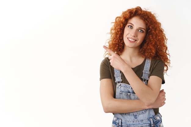 Mulher simpática e elegante ruiva caucasiana de cabelos cacheados convidar colegas de trabalho depois do trabalho, apontando o dedo para o espaço da cópia em branco, sorrindo despreocupada, mostrando uma proposta incrível, parede branca de pé