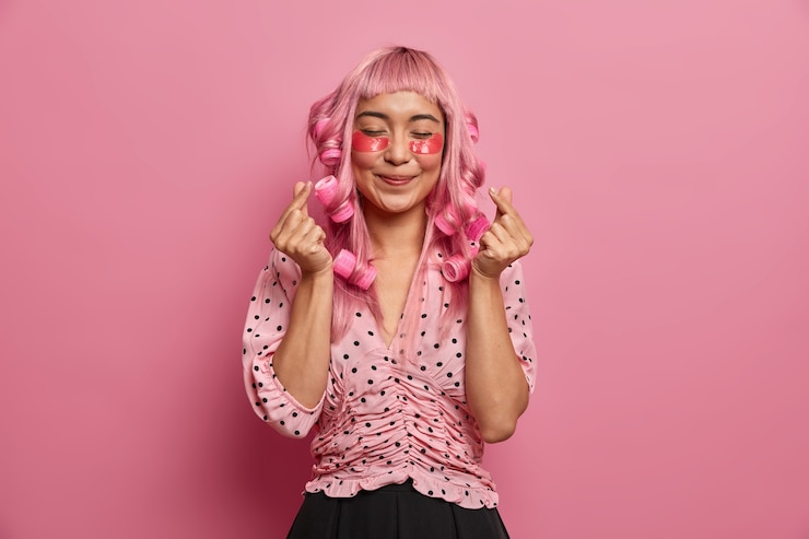 Foto grátis mulher sincera e positiva com longos cabelos rosa, passa rolos, faz penteado cacheado, mostra o sinal do coração coreano, usa manchas de colágeno rosado para reduzir o inchaço