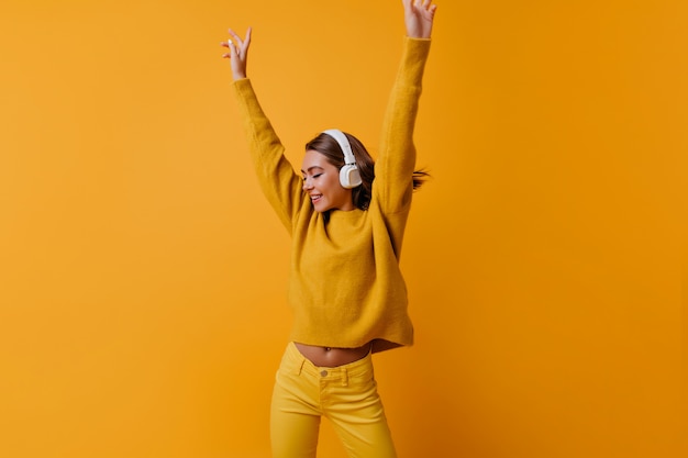 Mulher Slim positiva em calças amarelas dançando com as mãos para cima. Retrato interior da feliz garota em grandes fones de ouvido, desfrutando de uma boa música.