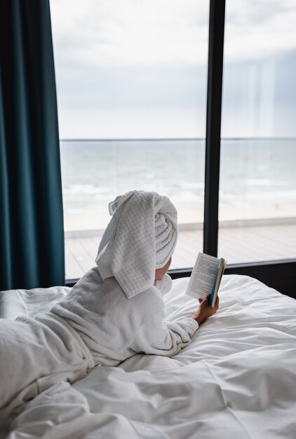 Mulher solitária com uma túnica branca e uma toalha deitada na cama e lendo um livro perto da janela