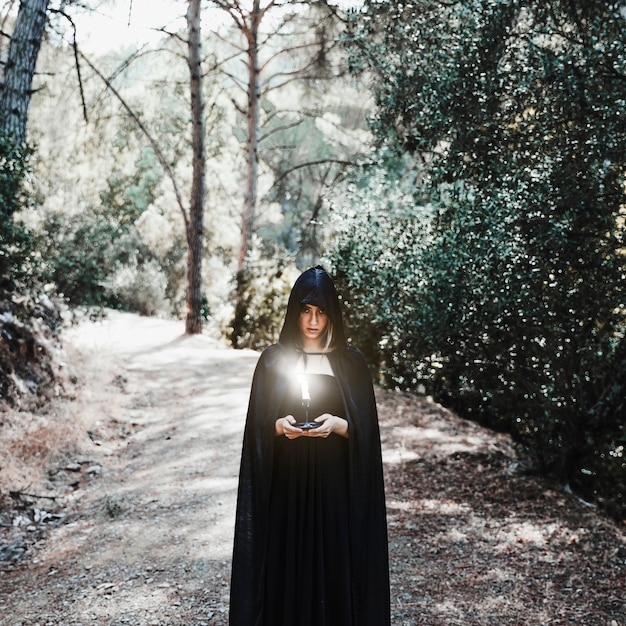 Foto grátis mulher sombria na capa segurando vela acesa na floresta ensolarada