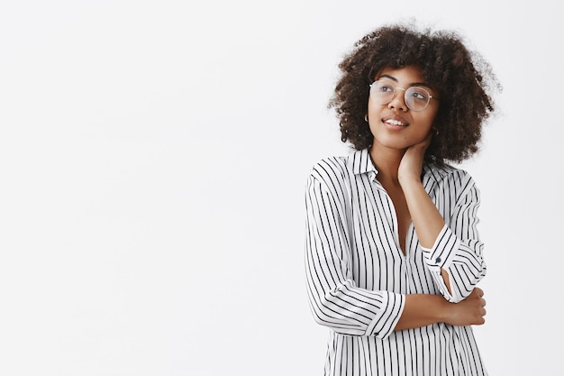 Mulher sonhadora e bonita, workaholic de pele escura, usando uma blusa listrada e óculos tocando o pescoço, sorrindo gentilmente e olhando para o canto superior esquerdo com uma expressão terna e romântica