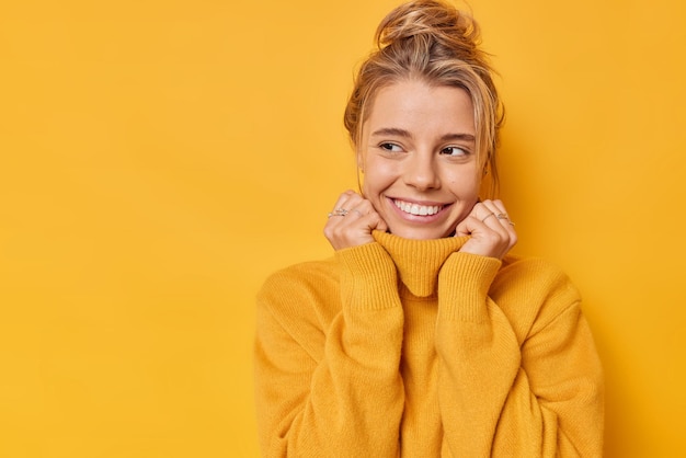 Mulher sonhadora positiva sorri suavemente desviando o olhar estando de bom humor usa um macacão quente e macio mantém as mãos no colarinho isolado sobre fundo amarelo com espaço de cópia em branco para seu conteúdo promocional