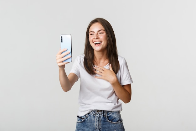 Foto grátis mulher sorridente amigável videochamada e falando no smartphone feliz, branco de pé.