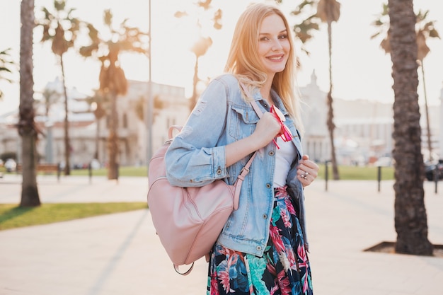 Mulher sorridente andando na rua da cidade com uma jaqueta jeans grande e elegante, segurando uma mochila de couro rosa