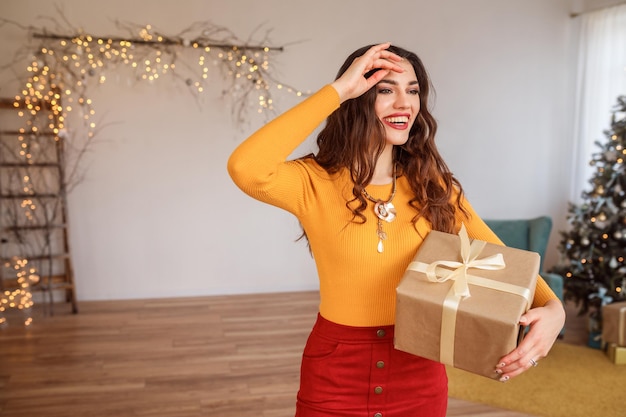 Mulher sorridente atraente com caixa de presente nas mãos, comemorando o natal em casa, no quarto festivamente decorado.