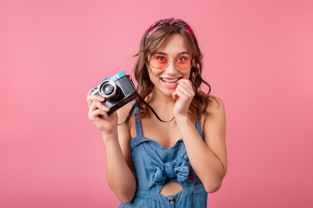 Mulher sorridente atraente com expressão emocional engraçada no rosto com câmera vintage em vestido jeans e óculos escuros isolados no fundo rosa