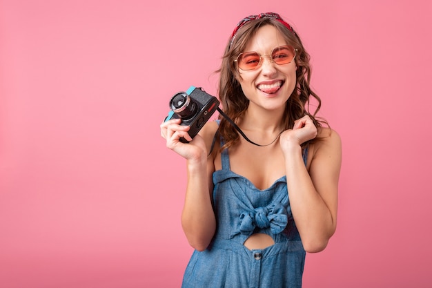 Mulher sorridente atraente com expressão emocional engraçada no rosto com câmera vintage em vestido jeans e óculos escuros isolados no fundo rosa