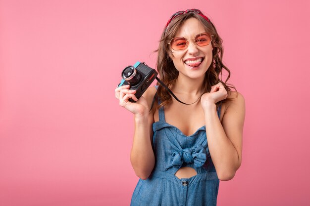 Mulher sorridente atraente com expressão emocional engraçada no rosto com câmera vintage em vestido jeans e óculos escuros isolados no fundo rosa