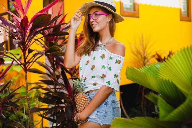 Mulher sorridente atraente de férias em t-shirt estampada com chapéu de palha na moda do verão, mãos segurando abacaxi