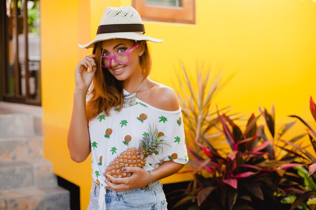 Mulher sorridente atraente de férias em t-shirt estampada com chapéu de palha na moda do verão, mãos segurando abacaxi