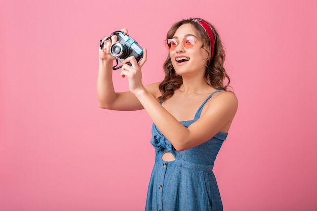 Mulher sorridente atraente tirando foto em uma câmera vintage usando vestido jeans e óculos escuros, isolado no fundo rosa