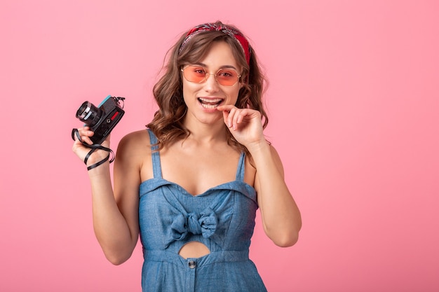 Mulher sorridente atraente tirando foto em uma câmera vintage usando vestido jeans e óculos escuros isolados no fundo rosa, expressão de rosto engraçado glamour