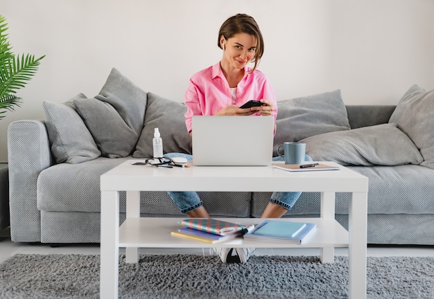 Foto grátis mulher sorridente com camisa rosa sentada relaxada no sofá em casa à mesa trabalhando online no laptop