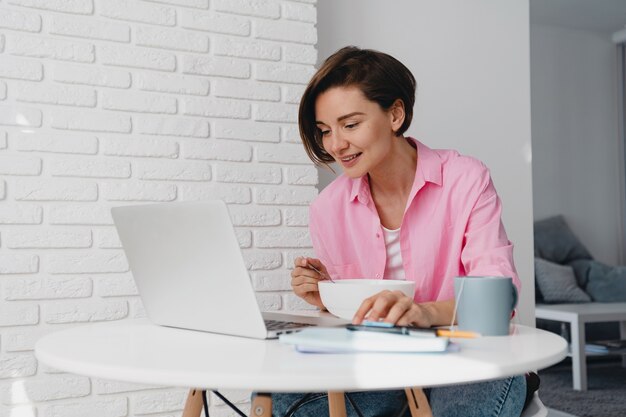 Mulher sorridente com camisa rosa tomando café da manhã em casa na mesa trabalhando online no laptop de casa, comendo cereais