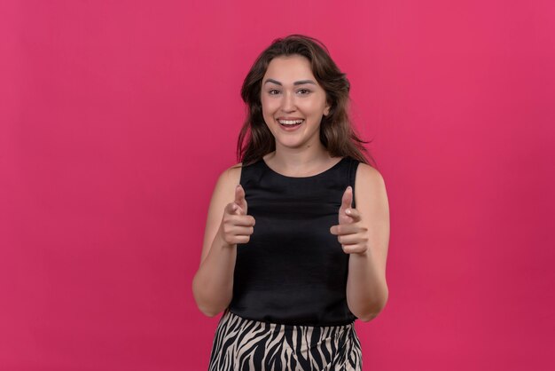 Mulher sorridente com camiseta preta apontando para frente na parede rosa