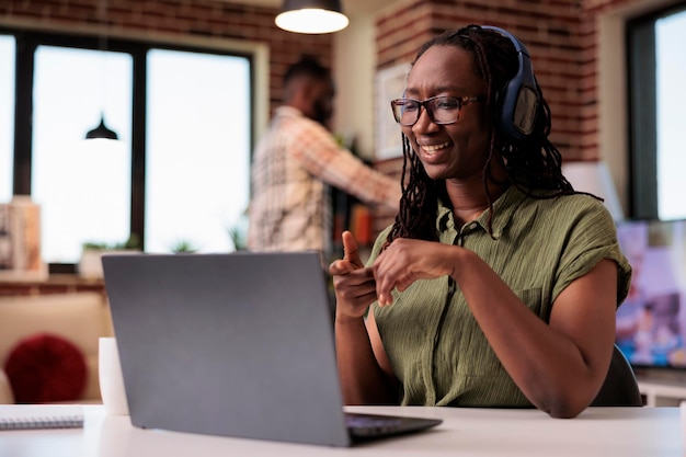 Foto grátis mulher sorridente com fones de ouvido sem fio assistindo show de entretenimento no laptop enquanto está sentado na mesa. freelancer afro-americano olhando conteúdo engraçado de mídia social em computador portátil.