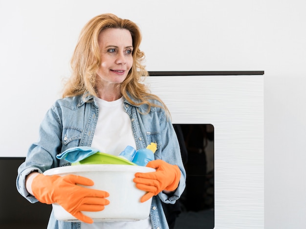 Mulher sorridente com luvas de borracha, segurando a cesta de produtos de limpeza