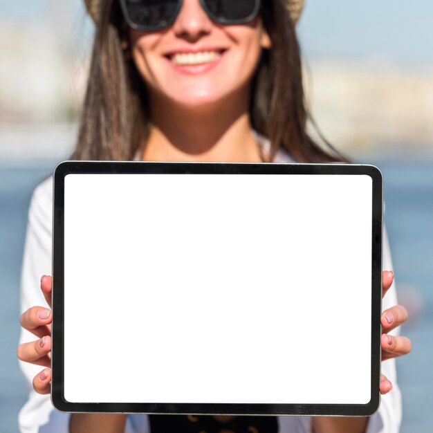 Mulher sorridente com óculos escuros segurando um tablet na praia