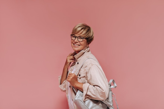 Mulher sorridente com penteado loiro elegante, óculos e bolsa cinza na jaqueta bege moderna, olhando para a câmera no pano de fundo rosa.