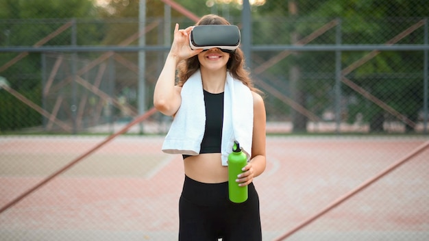 Foto grátis mulher sorridente com toalha usando fone de ouvido vr em um agasalho segurando uma garrafa de água em um campo esportivo