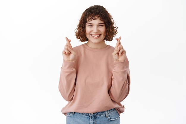 Mulher sorridente confiante cruza os dedos para dar sorte, fazendo o desejo parecer determinado e assertivo ao orar em pé contra um fundo branco copiar espaço