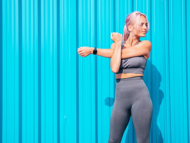 Mulher sorridente de fitness em roupas esportivas cinza com cabelo rosa Jovem bela modelo com corpo perfeitoFêmea posando na rua perto da parede azulAlegre e feliz Esticando-se antes do treino