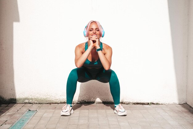 Mulher sorridente de fitness em roupas esportivas verdes com cabelo rosaEla fazendo agachamentos Jovem linda modelo com corpo perfeitoFêmea na rua perto de parede brancaAlegre e feliz Ao ar livre em fones de ouvido