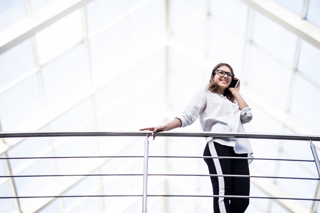 Mulher sorridente de negócios bem-sucedidos em pé, descansando e olhando através da varanda no moderno centro de escritórios, vestida com uma camiseta branca