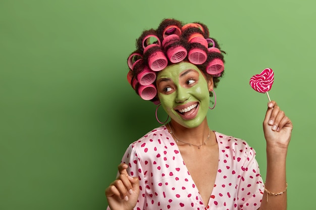 Mulher sorridente e alegre dança despreocupada, mexe-se com os braços erguidos, se diverte em casa, passa por procedimentos de beleza, aplica máscara facial para refrescar a pele, usa rolos de cabelo, segura pirulito delicioso