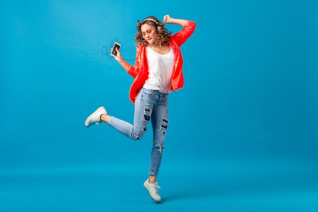 Mulher sorridente e atraente feliz dançando ouvindo música em fones de ouvido, vestida com roupa de estilo hipster isolada no fundo azul do estúdio, vestindo jaqueta rosa e óculos escuros