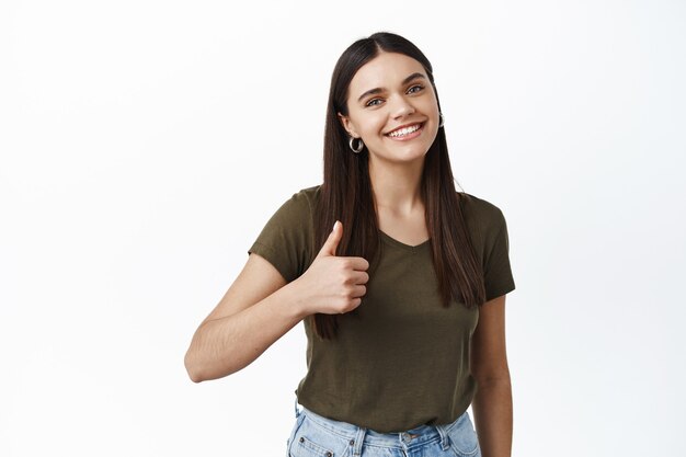 Mulher sorridente e atraente mostrando o polegar em aprovação, dar uma resposta positiva, aprovar ou elogiar algo, encostada em uma parede branca