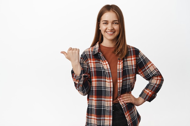Mulher sorridente e confiante apontando o polegar para a esquerda, mostrando o anúncio ao lado, anunciando, indicando no gráfico ou diagrama, em pé com roupas casuais contra a parede branca