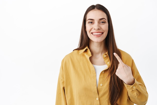 Mulher sorridente e confiante apontando para si mesma com um rosto orgulhoso e determinado, que se autopromove, mostrando seus novos dentes brancos, em pé sobre a parede do estúdio