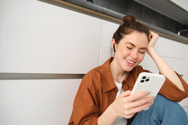 Mulher sorridente e despreocupada com mensagens de smartphone usando telefone celular sentada no chão da cozinha