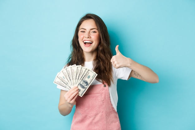 Mulher sorridente e feliz segurando dinheiro, notas de dólar e mostrando o polegar, recomendando um empréstimo de dinheiro rápido e parecendo satisfeita, em pé sobre um fundo azul