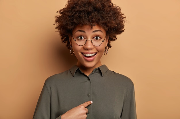 Foto grátis mulher sorridente e feliz surpreende que alguém cite seu nome, aponta para si mesma, não esperava ganhar ou ser escolhida, pergunta ao chefe se exatamente ela foi promovida, usa óculos redondos e camisa.