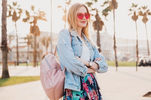 Mulher sorridente e flertando andando na rua da cidade com saia estampada elegante e jaqueta jeans grande e óculos de sol rosa