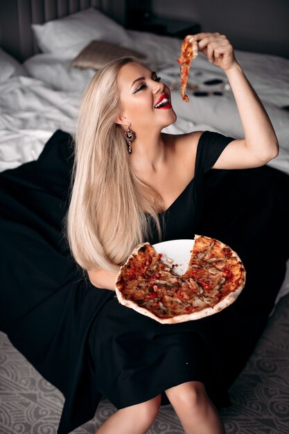 Mulher sorridente e muito elegante em um vestido preto segurando um pedaço de pizza enquanto está sentada na cama do apartamento dela